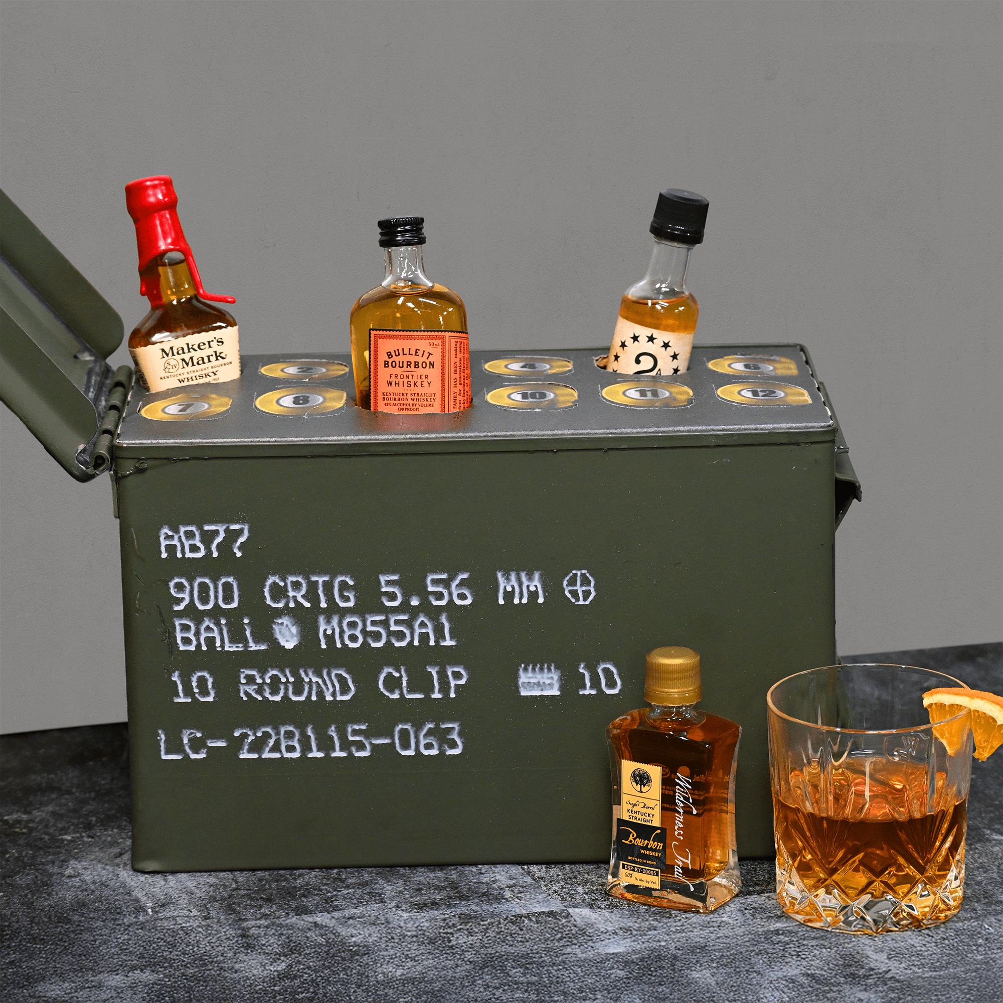 Ammunition can with mini bottles of whiskey and a glass on a gray surface